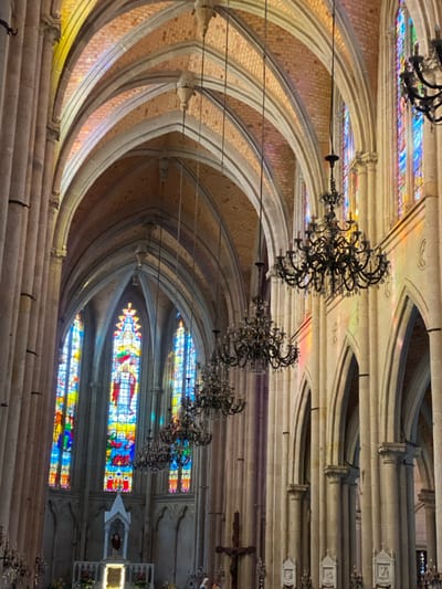 This image captures the majestic interior of a grand architectural space, likely a church or cathedral, viewed from a slightly elevated perspective towards the altar. The scene is dominated by intricate Gothic or Neo-Gothic architecture, featuring a series of high, pointed arches and ribbed vaulted ceilings constructed from light-colored stone and brickwork.

Numerous elaborate chandeliers, made of dark, ornate metal, hang in a line down the center of what appears to be the nave, extending into the distance. Along the right wall, several large, vibrant stained glass windows illuminate the interior with colorful patterns and depict religious figures and scenes. One particularly prominent window at eye level displays rich blues, reds, and yellows, illustrating a central figure.

Further down the right side, a small, white shrine or altar-like structure with a figure inside is visible. Above it, a dark wooden crucifix hangs on the wall. On the lower left of the image, long rows of stone or concrete benches, characteristic of church pews, extend into the foreground, suggesting a seating area. No people are present, and no specific activity or event is taking place, creating a serene and static ambiance.

The natural light streaming through the stained glass windows suggests it is daytime. The architectural style, stained glass, and religious artifacts strongly indicate a place of worship. Given the provided context, this is likely a prominent church in Guangzhou, China, such as the Sacred Heart Cathedral. No visible text is present in the image.