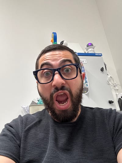A close-up, head-and-shoulders shot shows a person with a full beard and glasses, looking directly at the camera with a surprised or shocked expression. Their mouth is wide open in a gasp. The person is wearing a dark grey, textured long-sleeved shirt.

In the background, a white, flat surface, likely a wall or furniture, is visible. Several objects are placed on this surface. To the left, a small, white security camera with a purple light is visible, along with a blue and yellow toy or object. Further to the left, a blue vertical sign reads "HECON" in white lettering. Next to it, text that appears to be "R 2.4.1" is also visible. A miniature skateboard with a colorful graphic is propped against the surface. Wires are visible in the upper right portion of the background, suggesting an indoor setting, possibly an office or workspace.

The image appears to be taken indoors, with neutral, bright lighting. There are no strong indicators of the time of day or weather conditions outside. The overall mood is one of surprise or alarm, conveyed by the person's facial expression. The location context provided is Keila, Estonia.