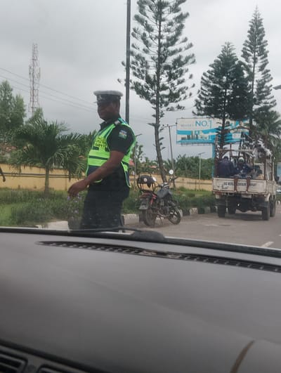 In Ikeja, Nigeria, a police officer in uniform stands by the roadside, wearing a high-visibility vest over a dark uniform. The vest is green and yellow, with the words "POLICE SPECIAL TRAFFIC" visible on it. He is wearing a hat and appears to be on duty, possibly directing traffic or conducting a checkpoint.
In the background, several tall pine trees are prominent, along with palm trees and lush green vegetation. A billboard advertising "Midea World's NO. 1" is visible in the distance, along with a cell tower. A white pickup truck with people and goods loaded in the back is stopped nearby. A motorcycle is parked on the side of the road. The sky is overcast, suggesting a cloudy or potentially rainy day. The scene appears to be a typical roadside setting in a Nigerian city.