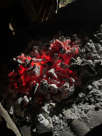 A close-up, high-angle shot focuses on a bed of glowing red-hot coals, partially covered by white ash, within a dark grilling pit. Several hands are visible in the upper portion of the frame, silhouetted against a dimly lit background, suggesting someone is tending to the coals. The scene is set outdoors at night, likely within the city of Durumi, Nigeria, a location known for its residential areas and informal markets where such grilling activities are common. The raw, intense glow of the coals contrasts with the surrounding darkness and the muted tones of the ash, creating a stark and primal visual. The texture of the ash and the irregular shapes of the coals are clearly defined, emphasizing the heat and potential for cooking.