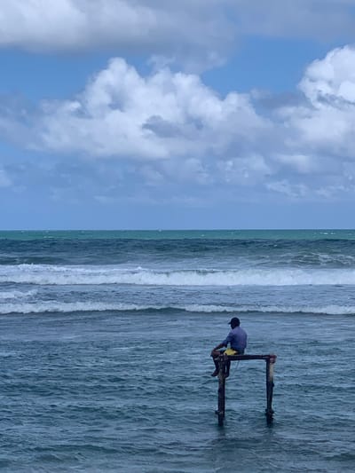 A lone man sits on a wooden structure submerged in the ocean. He is wearing a blue shirt, yellow shorts, and a dark baseball cap. The wooden structure consists of two vertical posts supporting a horizontal beam on which the man is seated. The ocean is choppy with waves breaking in the mid-ground. The horizon line is clearly visible with the sea meeting the sky. The sky is partially cloudy with a mix of blue sky and white cumulus clouds. The overall scene is one of solitude and contemplation by the sea. The location is identified as Kathaluwa, Sri Lanka.