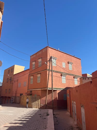 The image shows a street scene in Ouaouizert, Morocco. Tall, salmon-colored buildings line a narrow paved street. The buildings feature rectangular windows with white shutters. Utility poles and power lines are visible. A person walks on the right side of the street, casting a shadow. The sky is clear and blue. A woven bamboo-like material is attached to the side of one building.