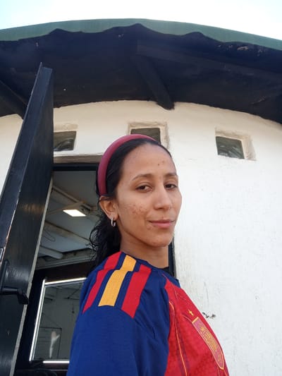 A medium shot photograph, taken from a low angle, shows a young woman looking towards the camera. She is wearing a red headband, a hoop earring in her left ear, and a blue shirt with red and yellow stripes across the shoulders. The shirt also has a red sleeve with a gold crest and yellow checkered pattern. The woman has medium-length, dark hair that is wet and appears to be pulled back from her face. Her skin is light brown with some freckles and blemishes visible. Her lips are slightly parted, and she has a neutral expression.
She is standing in front of a white, textured building with a dark, sloped roof. Two small, square windows are visible on the building's surface above and to the right of the woman's head. To the left of the woman, a dark-colored, partially open door is visible, revealing a glimpse of an interior space with a fluorescent light fixture. The architectural style suggests a structure possibly in a tropical or arid climate, consistent with locations like Porlamar, Venezuela, known for its warm weather and distinctive coastal buildings.