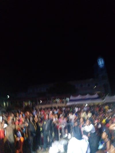 A large crowd gathers at night, seemingly at an outdoor event. People are dressed in varied colors and styles. In the background, a building with a prominent tower is faintly visible, suggesting a possible landmark. A blue and white striped awning sits above the crowd. The overall image appears blurry and low in resolution.