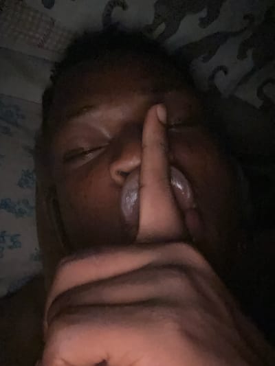 The image shows a close-up of a person's face, with their eyes closed and a finger pressed to their lips, in a gesture of silence. The person has dark skin and is lying down, possibly in bed, as indicated by the patterned fabric in the background which appears to be a duvet or bedsheet. The lighting is dim, suggesting it might be nighttime or a dark room. There is no specific information to determine the location as Ikorodu, Nigeria.