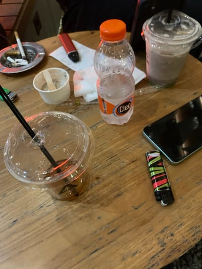 The image shows a wooden table with a variety of objects on it, including a plastic cup with a black straw, a plastic cup containing what appears to be iced coffee, a bottle of water, a small plastic cup, a lighter, a black cell phone, a red and black vaporizer, a pack of cigarettes in a plastic ashtray, and a white napkin. The table is made of wood with a distinct grain pattern. The location is Malang, Indonesia, based on the provided coordinates. The table is situated in an outdoor setting with a slight blur of grass or other greenery visible in the background. The image is taken from a bird's-eye perspective, looking down on the table and its contents. The table appears to be a typical outdoor table for a cafe or restaurant. The objects on the table suggest that people have been sitting at the table recently. The cell phone, lighter, and vaporizer suggest that the people were perhaps enjoying a break or a social gathering. The image depicts a typical scene of a cafe or restaurant setting in Malang, Indonesia.