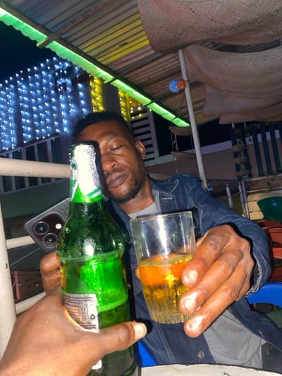 A man is reclined, eyes closed, while wearing a denim jacket over a gray shirt. He holds a partially filled glass of amber liquid above a green bottle. His phone rests underneath the bottle. In the background, there's a thatched roof with decorations and a fence illuminated by strings of white lights. The shot is taken at an angle with the scene partially obscured.