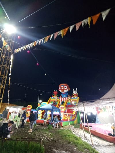 This image depicts a nighttime scene at an outdoor fairground or temporary entertainment area in Khe Sanh Commune, Vietnam. The central feature is a large, multi-colored inflatable play structure adorned with cartoon characters, including a prominent Pororo the penguin figure at the top, a red frog, and a white rabbit. Several individuals, including children and adults, are visible around and on the inflatable.
To the left of the inflatable, a vendor stall with a light-colored canopy is present, with several individuals gathered near it. In front of the inflatable, a small blue canopy tent shelters two seated individuals. To the right, a large, white tarp-like tent structure is supported by wooden poles, with pink fabric or inflatable elements visible beneath it.
The scene is illuminated by multiple artificial light sources. A tall metal lattice tower on the far left emits a bright white light, causing lens flare. Strings of triangular pennant flags in orange, yellow, white, and pink are strung across the upper portion of the frame, along with several smaller, spherical colored lights (red, green, yellow) hanging from wires.
Approximately 10-15 individuals, mostly adults and teenagers, are present, casually dressed. One person in the immediate foreground, wearing a dark jacket, is bent over looking downwards. The ground is uneven, composed of dirt and sparse grass, suggesting an undeveloped or temporarily utilized space. The sky is dark, indicating night, with a faint light source or reflection visible in the upper right.