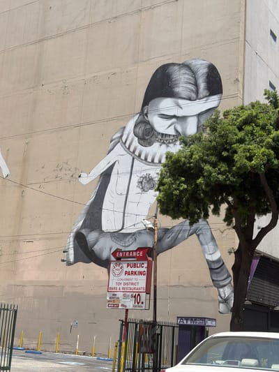This is a photograph of a mural on the side of a building in Los Angeles, California, United States of America. The mural is a black and white illustration of a large figure with its eyes covered by its hands. The figure is wearing a collared shirt and striped pants. The mural covers a significant portion of the building's facade. In front of the building, there is a tree with green foliage. There is also a street sign that reads "ENTRANCE PUBLIC PARKING" and lists parking rates. A white car is parked in the foreground, partially obscuring a fence. The scene appears to be in an urban environment, possibly a commercial or entertainment district, given the "TOY DISTRICT BARS & RESTAURANTS" text on the parking sign. The lighting suggests it is daytime.