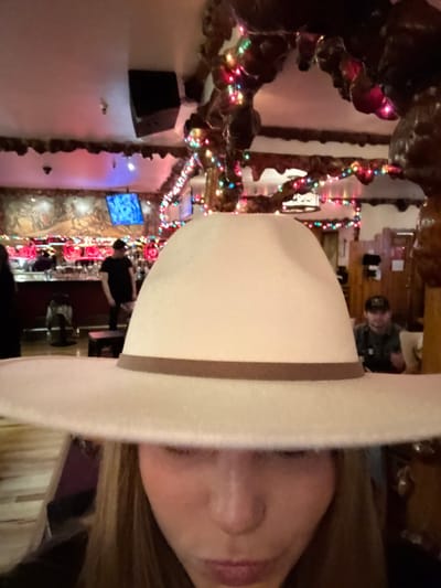 A woman wearing a large white cowboy hat dominates the frame. Behind her, a restaurant interior is visible, decorated with Christmas lights along the walls and archways. A television screen glows blue amidst the decorations. Other patrons can be seen at the bar in the background.