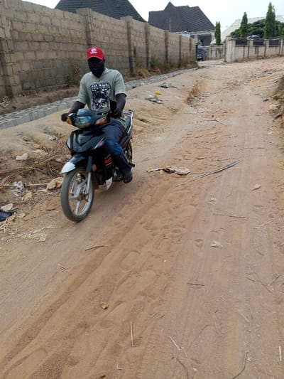 A person wearing a red cap rides a black motorcycle on a dirt road. The road is unpaved and shows signs of wear with scattered debris. A partially constructed brick wall runs along the left side of the road. In the background, houses with dark roofs and a few trees are visible under an overcast sky. The overall scene suggests a rural or developing area.