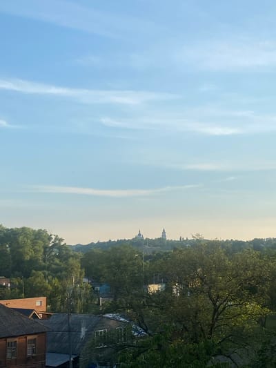 The image depicts a panoramic view of the city of Chernihiv, Ukraine, from an elevated perspective. The sky is a clear, pale blue with thin, wispy clouds scattered across it. In the distance, on a hill overlooking the city, are several domed structures, likely churches or cathedrals, characteristic of Ukrainian architecture. The midground is a dense expanse of green trees, with a scattering of rooftops of houses visible amongst the foliage. The foreground features several older residential buildings with dark, sloped roofs. The overall impression is one of a peaceful, rural or semi-rural setting within a city, bathed in the soft light of what appears to be late afternoon or early morning. There are no people visible in the image, and no discernable actions or interactions are taking place. The scene is static and serene.