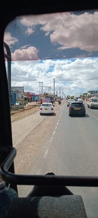 A street view shows cars moving on a paved road with a broken white center line. Buildings and signs line the left side of the road. Power poles with wires extend along the road's edge. The sky is visible through a partially tinted window, reflecting clouds. The image is taken from within a vehicle, capturing parts of the interior, including a seat and frame.