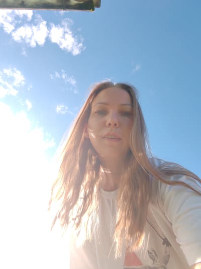 An over-the-shoulder, low-angle selfie taken outdoors under a bright blue sky with scattered white clouds. The sun is positioned in the upper left of the frame, creating a strong lens flare that washes out much of the left side of the image and illuminates the subject's hair.
The subject is a woman with long, light brown hair, fair skin, and light-colored eyes. She is wearing a white crew-neck t-shirt with a graphic print on the lower right side, partially obscured by the sun's glare. Her facial features are visible, with her mouth slightly open. The camera angle looks up at her from a position below and in front of her.
In the upper left corner of the image, a portion of a wooden structure, possibly a roof or overhang, is visible. The overall scene suggests a sunny day, likely during daylight hours. The location is specified as Chuchul'ma, Russia.