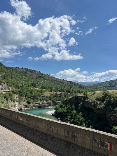The image depicts a view from a bridge in Donje Mrke, Montenegro. The sky is blue with white, puffy clouds. Below the bridge, a turquoise river flows through a forested valley. The bridge's concrete railing is visible in the foreground. A red and white trail marker is affixed to the railing. Vegetation covers the steep hillsides surrounding the river.