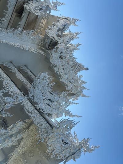 A low-angle photograph captures an ornate section of the Wat Rong Khun (White Temple) located in Ban Rong Khun, Thailand. The architectural details consist of intricately sculpted, pure white plaster or concrete adorned with numerous embedded reflective mirror fragments, which glitter under direct sunlight. Visible elements include multi-tiered rooflines, decorative gables, and elaborate finials with traditional Thai mythological motifs. The bright white structure occupies the left and bottom portions of the frame, sharply contrasting with a clear, vivid blue sky that dominates the upper right. A solitary wisp of white cloud is visible in the upper right quadrant of the sky. No individuals, actions, or interactions are depicted in the scene.