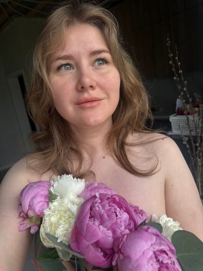The image is a close-up portrait of a young woman, with the photograph rotated 90 degrees counter-clockwise. The woman appears to be lying down or reclining, looking towards the camera with a serene and softly smiling expression. She has light-colored, wavy hair (blonde or light brown) that frames her face and flows over her shoulders; a distinct strand on her bare shoulder forms a heart shape. Her fair skin features natural freckles and a few moles, and she has striking light blue eyes.
A vibrant bouquet of flowers rests on her chest and shoulder area, partially covering her upper body. The arrangement is dominated by large, lush pink peonies and smaller, ruffled white flowers, possibly carnations, accented with green foliage, likely eucalyptus leaves. Both the woman's face and the flowers are in sharp focus.
The setting appears to be indoors, with a dark, blurred, and out-of-focus background creating a shallow depth of field. Subtle details in the background include fuzzy pussy willow branches, suggesting a springtime atmosphere. The lighting is soft and diffused, contributing to a gentle and intimate mood. There is no visible text in the image. The scene captures a moment of calm and natural beauty, located in an indoor setting in Ферма, Russia.