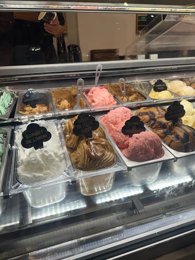 This image depicts the interior of a gelato shop, specifically focusing on an illuminated display case filled with various gelato flavors, presumably located in Acassuso, Argentina.
The central subject is a refrigerated display case containing multiple stainless steel and clear plastic tubs of gelato. Prominently visible flavors include "CHOCOTORTA" (a brown, textured gelato), "DULCE DE LECHE SÚPER" (a swirled light and dark brown gelato with a glossy top), "SORBET DE LIMÓN" (white, fluffy lemon sorbet), "SORBET DE FRUTILLA" (pink strawberry sorbet), "CHOCOLATE PASION" (dark brown gelato), and "DULCE DE LECHE GRANIZADO" (a creamy brown gelato with darker chocolate chips or swirls). Other tubs contain varying shades of pink, cream, and brown gelato, some with visible textures or inclusions. Each labeled flavor has a black, cloud-shaped marker with white text. Metal serving spoons are present in several tubs.
Above the gelato display, a reflective surface shows the blurred reflection of an individual, likely a staff member or customer, holding a small dark cup with a white design in their right hand. A smartphone, possibly a black iPhone, is also visible in their left hand, held up as if taking a photograph. The background reflection suggests a clean, minimalist interior with white walls, a counter, and stacked brown trays. The lower section of the display case features linear LED lighting, illuminating the gelato from below. No direct interactions or actions are clearly observable beyond the reflection.