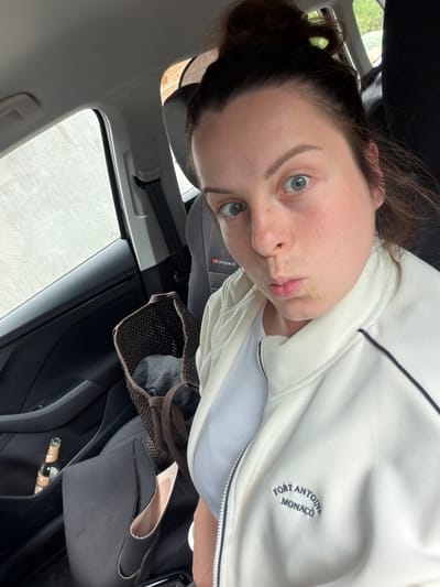 A woman is sitting in the passenger seat of a car, looking at the camera with her lips pursed. She has her hair pulled up in a bun and is wearing a white zip-up jacket over a light blue top. A brown mesh bag with some dark fabric inside is in the backseat. A bottle of drink is visible in the door pocket on the left. The interior of the car, including the dashboard and window, is visible. The image appears to be taken in Gangloff, Germany.