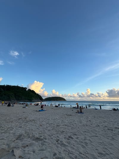 The image depicts a crowded beach scene in Rawai, Thailand, likely during sunset given the lighting. The foreground is dominated by sand, with footprints and some scattered debris. In the midground, numerous people are seated or lying on the sand, some on towels or mats, and others are walking or standing. The middle ground also shows the edge of the ocean with gentle waves, and several individuals are in the water, some with surfboards.
Further back, a lush green, tree-covered hill forms the left side of the scene, with a small island visible in the distance to the right. The sky is a vibrant blue with scattered, puffy white clouds, some illuminated by the setting sun. The overall impression is a lively, recreational atmosphere on a tropical beach.