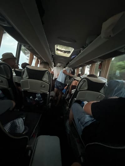 The image depicts the interior of a bus, likely during a trip in Ca' di Valle, Italy. Several passengers are seated in upholstered chairs. The aisle is visible between the rows of seats. Overhead compartments are present above the seating area. Outside the windows, glimpses of trees and buildings are visible. Water bottles are stowed in seat pockets.