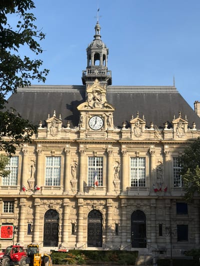 This media file depicts the exterior of a historical building, likely a town hall, in Ivry-sur-Seine, France. The building features ornate architecture with stone carvings and multiple stories of windows. A prominent clock face is visible on the facade. To the left of the main building is a tall, dark tower with a spire and weather vane. The sky is clear and blue, suggesting a sunny day, likely in the morning or afternoon given the lighting.

Several flags, appearing to be French tricolor, are affixed to the building. There are also inscribed words above doorways, including "FRATERNITE" and "LIBERTE", which are significant French values. In the foreground, on the right side of the image, several construction vehicles are parked, including a red tractor and two yellow excavators. Trees and foliage surround the building, indicating an outdoor setting within a town or city. No people are clearly visible, but the presence of construction equipment suggests ongoing work or recent activity.