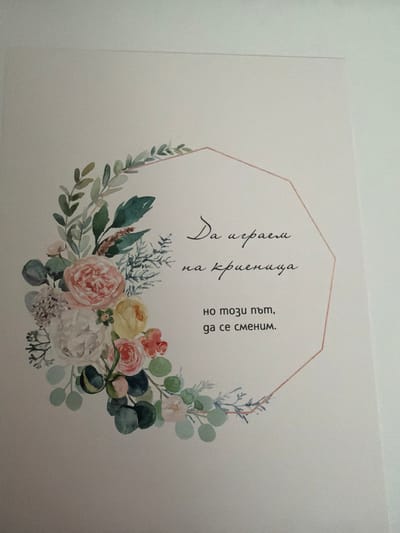 This is a close-up, eye-level, indoor photograph of a piece of paper with a floral watercolor design and text. The paper appears to be a card or a print. A delicate border of watercolor flowers and greenery, predominantly in shades of pink, white, yellow, and green, frames the left and top edges of the paper. A polygonal shape, outlined in a light peach color, is present in the center right of the image. Within this shape, there are three lines of handwritten text in Bulgarian. The text appears to be a quote or a message. The background is a plain, light-colored surface, likely a wall or a table, with subtle texture and slight shadowing indicating indoor lighting. There are no people or animals visible in the image. The photograph is taken with a slight angle, and the lighting casts subtle shadows. There is no information in the image that would indicate a specific location in the city of Pazardzhik, Bulgaria.