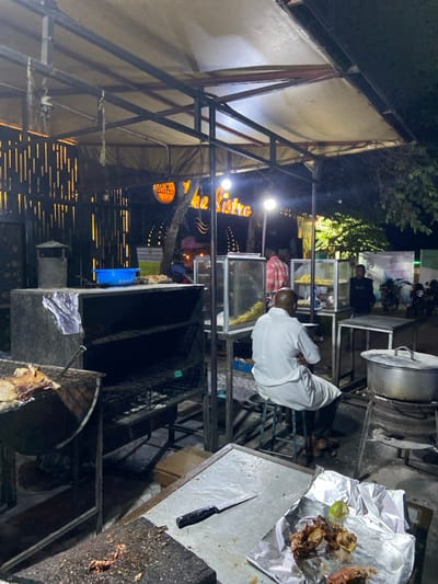 The image shows an outdoor food stall in Dodoma, Tanzania, located at the coordinates -6.157 latitude and 35.723 longitude. The stall is situated under a canopy, with a sign that reads "The Bistro" illuminated with warm yellow light. A man in a white shirt sits on a stool facing away from the camera, with his back to the viewer. The stall appears to be a barbecue restaurant with a large metal grill, where food is being cooked over an open flame. There are other food stalls nearby, selling snacks and drinks. The image captures a dimly lit night scene, with the light from the stalls illuminating the space. The ground is uneven, with a mixture of pavement and dirt. A large metal pot sits on a stand in the foreground, with a small knife resting on the table. A plate covered in foil holds food, likely prepared for a customer.