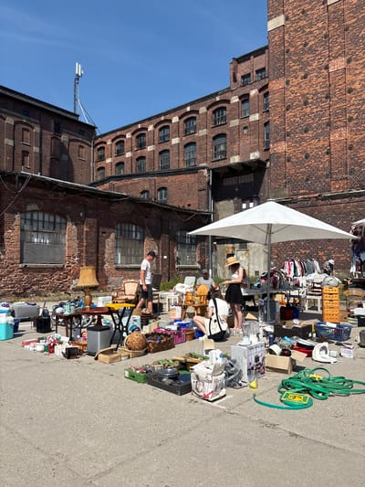 This is a ground-level, wide shot of an outdoor flea market in Wrocław, Poland. The scene is dominated by a large brick industrial building complex, which provides a weathered backdrop. The ground is asphalt.
In the foreground, a variety of goods are laid out on the ground and tables. These include household items such as appliances (a blender, a fan), kitchenware (pots, pans, mugs), lamps, boxes, baskets, and cleaning supplies (a green garden hose). A bicycle is also visible among the items.
Several people are present. A man in a white t-shirt and black shorts is walking through the market. A woman in a straw hat and a black skirt, along with another woman in a white top and shorts, are browsing near a white umbrella. A man is sitting on a chair in the background. Clothing is displayed on a rack near the umbrella.
The sky is clear and blue, indicating a sunny day. A cell tower is visible on top of one of the buildings. The overall atmosphere is that of a typical weekend flea market with vendors and customers interacting amidst a collection of used goods.