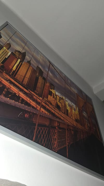 The image is a photograph of a large framed artwork depicting the Brooklyn Bridge and the Manhattan skyline at sunset. The artwork is hung on a white wall at an angle, with a section of the ceiling visible in the upper right corner. The artwork itself is a panoramic view, with the bridge's iconic cables and structure prominently featured in the foreground. The skyscrapers of Manhattan are visible in the background, illuminated by the warm glow of the setting sun. The colors are rich and vibrant, with shades of orange, yellow, and purple in the sky, and the warm tones of the buildings. The framing of the artwork appears to be a simple silver metal. The image does not contain any people or specific details related to the city of Kutná Hora, Czechia.