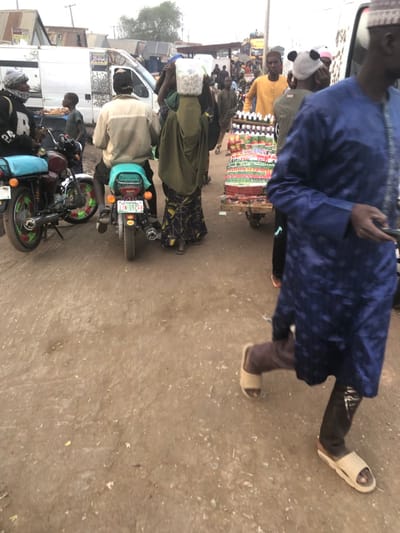 The image captures an unpaved, active street scene in Kufena, Nigeria, under diffuse daylight. The ground consists of compacted dirt with scattered debris.
In the immediate foreground, on the right, a male subject is partially visible, walking left-to-right. He wears a blue, patterned, long-sleeved garment over dark trousers and light-colored slide sandals, holding a dark object in his right hand.
In the mid-ground, a male subject is seated on a parked motorcycle, facing away from the camera. The motorcycle features a light green seat cover and a visible Nigerian license plate: 'KADUNA LJN-152QA'. To his left, another motorcycle is parked, distinguished by green and red patterned wheel rims and a light blue seat cover. A male subject wearing a dark hoodie stands near this second motorcycle. Directly in front of the seated motorcyclist, a female subject walks away from the camera, carrying a clear plastic container filled with small white objects balanced on her head. She is attired in a green head covering and a patterned long skirt.
Adjacent to the woman, a wooden vendor's cart is present, heavily loaded with multiple tiers of bottled beverages (soft drinks and water) and various packaged snacks. Behind this cart, several individuals are visible, including a man in a yellow shirt and another wearing a distinctive panda-ear beanie. The background reveals a crowded street, featuring a white panel van, other vehicles, and rudimentary structures consistent with a market environment, indicating high pedestrian traffic.