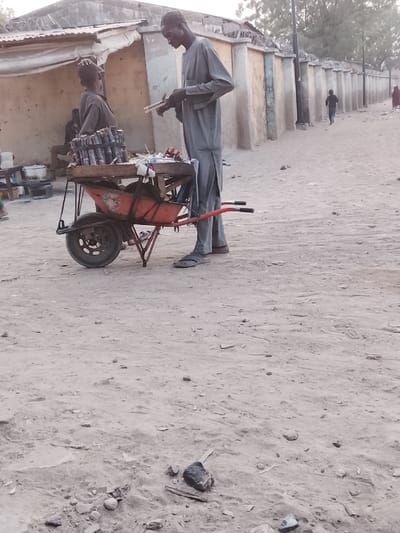 Two men stand on a dusty road, one tall, the other shorter. The taller man, dressed in a grey robe, holds small objects. Both are near a rusty orange wheelbarrow overflowing with canned drinks and items. In the background, there is a fence, buildings, and a person walking away. The ground is arid and uneven, with scattered rocks and debris.