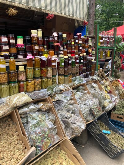 The image shows a market stall in Gyulagarak, Armenia, laden with various products. On the left, there's a tiered display of jars filled with preserves, jams, and pickled items. To the right are cardboard boxes and plastic crates containing dried herbs and herbal mixtures, some in clear plastic bags. Watermelons and bottles of liquids are visible on a higher shelf. A striped awning partially covers the left side of the stall.