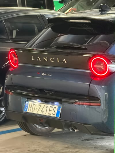 The image is an outdoor, low-angle shot of the rear of a dark gray Lancia Ypsilon. The car is parked on a concrete surface, with a light blue line visible near the bottom right of the frame, suggesting a parking spot. The scene appears to be a parking lot or garage, with no discernible weather or time of day cues.

The car's taillights are illuminated, one with a prominent red glow. The "LANCIA" branding is visible in silver lettering on the car's rear, along with the "Ypsilon" model name. A license plate is attached to the rear of the car, displaying the text "HD-741ES". The overall impression is a focused view on the car's design and details. No people are visible in the image.