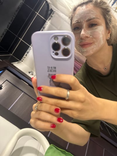 The image shows a young woman in a bathroom, taking a selfie with her phone. She has a white facial mask on her face, and her eyes are open, looking at the camera. Her hair is blonde and tied back. She is wearing a dark green t-shirt. The bathroom has black and white tiled walls, and dark wood cabinets. A white sink and a white towel rack are also visible. The woman is wearing a silver ring on her left hand, which is holding the phone. Her fingernails are painted red. There is some text visible on the back of the phone case, which appears to be an illustration of a cat and some Russian text. The lighting suggests it is daytime, and the environment appears to be indoors in a residential setting. The woman's expression is neutral, perhaps a bit posed for the selfie. The overall impression is of a casual moment of self-care.