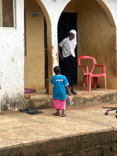 An outdoor scene depicts two females in Ikorodu, Nigeria. The foreground shows a young female wearing a blue t-shirt with the inscription "AYOMIDE" and a pink skirt, holding a plastic water bottle. She is standing on a concrete surface with a few dark clothes on the ground behind her. To her right, an adult female wearing a white hijab and a striped t-shirt over a black skirt stands near a bright red plastic chair. The background features an arched entrance to a building with yellow walls, and part of a white exterior wall. The environment appears to be a residential area with a slightly weathered appearance.
