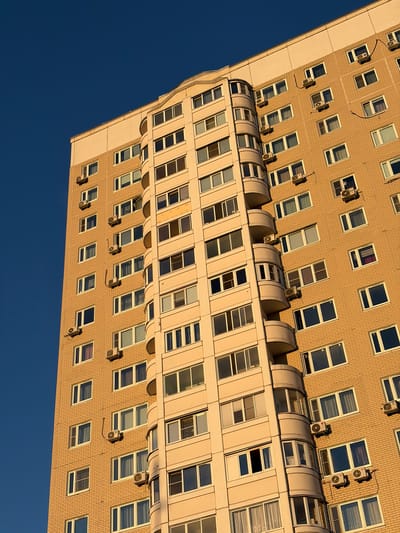 This is a downward-facing, low-angle shot of a modern, multi-story building, viewed against a clear, deep blue sky. The building is clad in light brown brickwork and features numerous windows, many of which are large and rectangular. Several windows have distinct blinds or curtains visible within them, suggesting interior spaces.
Prominently attached to the exterior of the building are numerous air conditioning units, mounted in various configurations next to the windows. These units are a recurring detail throughout the visible facade. The building's design incorporates a series of curved, translucent roof structures that appear to cover balconies or access points. Below these, another set of windows is visible, featuring a lighter-colored frame and a different architectural style.
The perspective and the sharp shadows cast by the air conditioning units indicate that the sun is relatively low in the sky, suggesting it is either early morning or late afternoon. The clear sky implies fair weather. The context provided places the location in Moskovskiy, Russia. There is no visible text in the image. The overall impression is one of urban architecture and a functional, lived-in building.