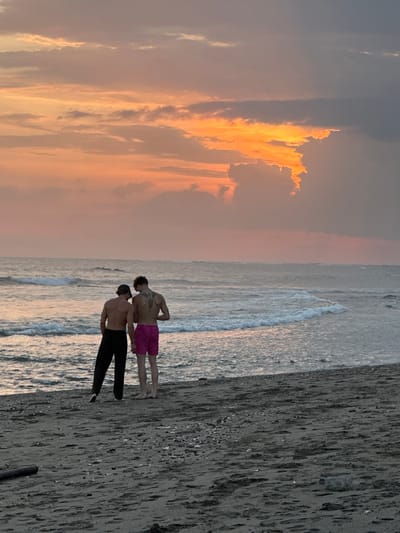 Two shirtless young men stand on a sandy beach, facing the ocean, during sunset. The man on the left wears black pants and a dark baseball cap. The man on the right wears bright pink shorts and has a large tattoo on his back. Waves gently break on the shore, and the sky is a mix of orange, pink, and gray clouds, illuminated by the setting sun. The beach is dark sand with scattered pebbles and debris. The scene is likely located on the coast of Mengwi, Indonesia, a region known for its beaches and sunsets.