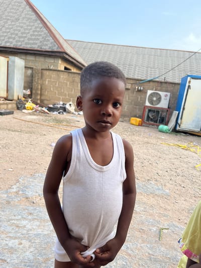 A young child wearing a white undershirt stands outside in Iddah, Nigeria. The location appears to be near or behind buildings made of concrete blocks. There is debris scattered on the ground, and an air conditioning unit is visible on one of the buildings. The child is looking toward the camera. The sky is visible in the top left of the frame.