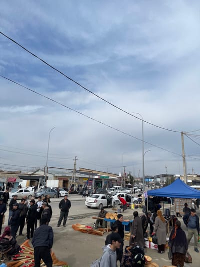 The image captures a wide-angle view of a busy outdoor market and street scene in Yangirabаt, Uzbekistan, under an overcast sky. The upper third of the frame shows a light blue-grey sky with scattered stratiform clouds, bisected by multiple diagonal overhead utility lines.
The central and lower portions of the image depict a multi-lane asphalt road extending horizontally across the midground, with vehicular traffic and numerous parked cars, predominantly white and silver sedans. Several vehicles have doors open. Commercial buildings line both sides of the street in the background, featuring storefronts, some with "DORITONA" branding visible in Latin script, and others displaying Cyrillic text. Utility poles with multiple wires and modern streetlights are dispersed along the roadside.
In the foreground and midground, dozens of individuals are present, engaged in various activities. Most people are dressed in casual cool-weather attire, including jackets, coats, and some hats. On the right side of the frame, a large blue canopy tent serves as a market stall, under which merchandise is displayed and shoppers are observed. Immediately in front of the blue tent, several individuals are standing and examining goods, some carrying bags. In the central foreground, bundles of brooms with red and brown bristles are laid out on the pavement for sale. Near these brooms, a male vendor sits on a stool beside a display of round, flat bread loaves. Other individuals are standing in groups, walking, and interacting along the paved areas adjacent to the street. The scene indicates active commerce and pedestrian movement within an urban commercial district.