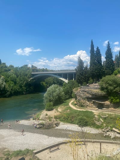 This is a horizontal, eye-level, medium shot of the Morača river in Podgorica, Montenegro on a sunny day. A modern, concrete bridge spans the river in the background. On the riverbank to the right, there are steep, rocky cliffs with some ancient ruins and a path leading up. Many people are visible on the rocky riverbed and shallow water of the Morača river, some are swimming, some are sitting or lying down on the shore. The river is a vibrant blue-green color. Lush green trees and vegetation are abundant along the riverbanks and on the cliffs. The sky is bright blue with scattered white clouds. A wooden bench is visible in the foreground on a paved area.