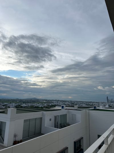 The image captures a view of Santa Clara Ocoyucan, Mexico, from a rooftop perspective. The sky is overcast with a mix of gray clouds and patches of blue, with sunlight breaking through in some areas. Below the sky, a sprawling cityscape is visible, characterized by numerous low-rise buildings with white facades and some green rooftops. In the distance, a mountain range can be seen. In the foreground, the white structures of what appear to be modern buildings with balconies and glass windows are prominent. A small, bare-branched tree in a pot sits on one of the balconies. The scene suggests an urban or suburban environment with residential or commercial architecture. There are no people or animals visible in the image. The lighting indicates it is likely daytime, possibly late afternoon or early morning, given the diffused light.