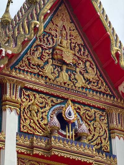 This is a close-up, eye-level shot of the exterior facade of a temple in Chalong, Thailand. The architecture is highly ornate, featuring intricate gold detailing and vibrant red roof elements. At the apex of the gable, a golden statue of Buddha is seated. Below this, a row of smaller golden figures, possibly monks or deities, are depicted in various postures. Decorative scrollwork, also in gold, adorns the facade. A band of Thai script is visible across the upper section of the facade, beneath the row of figures. Below this, a decorative archway frames a central ornament with a teardrop shape, surrounded by patterned tiles and small golden pillars. The entire structure appears to be adorned with mosaic tiles in shades of blue, red, and gold. The sky is a pale, overcast white.