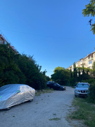 The image depicts a street scene in Sevastopol, Ukraine. On the left, a silver car is covered with a reflective tarp. To the right of it, a black sedan is parked. Further down the road, a silver SUV is parked on the right side. Behind the cars, there are multi-story apartment buildings and dense greenery. The sky is clear and blue.