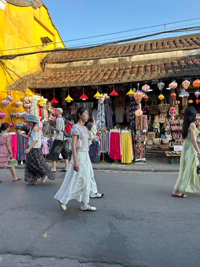 In a street scene in Hoi An Ward, Vietnam, several people walk past a brightly lit shop displaying a vibrant array of goods, including clothing and lanterns. The shop's facade is painted a striking yellow, with a tiled roof and an overhang draped with colorful lanterns of various shapes and sizes. In the foreground, a woman in a flowing light green dress walks to the right, her side profile visible as she holds a dark handbag. To her left, a woman in a white top and long white skirt walks towards the right, her hand raised slightly. Further left, another woman in a blue hat and plaid skirt walks to the right, looking at her phone. A man in a light-colored patterned shirt and dark shorts is also visible, walking to the right with a red backpack. A woman in a pink dress is seen on the far left, walking to the right with a fan. The street is paved with asphalt, and the overall atmosphere suggests a bustling market or shopping area in the city.