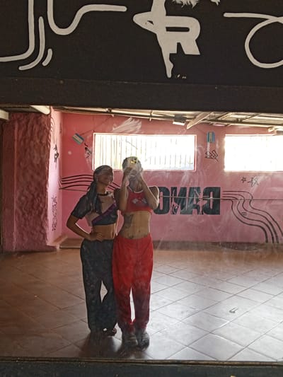 This image depicts two young women posing for a mirror selfie inside a dance studio located in El Espinal, Venezuela. The scene is dominated by the large mirror reflecting the studio's interior and the individuals.
The woman on the right is holding a smartphone, obscuring her face as she takes the picture. She wears a red crop top with a checkered pattern on the straps and loose red pants. Tattoos are visible on her stomach and arm. The woman on the left, with her face partially visible and a faint smile, wears a dark-colored crop top with some lighter accents and dark patterned pants. Her hair is tied back. Both are dressed in casual, athletic-style clothing suitable for dance.
The studio has light pink-colored walls and a light brown tiled floor. Bright daylight streams in through two large windows at the back. The walls are decorated with painted elements, including abstract black lines resembling sound waves or pathways, and white star decals. Reflected on the back wall, the word "DANCE" is clearly visible in large, dark, mirrored capital letters, confirming the studio's function. A small blue and white striped object, possibly a flag or part of one, is also affixed to the wall. Above the main reflection, the mirror shows part of a black surface with white, stylized text or artwork, including what appears to be a large, angular "R" and some curved, calligraphic shapes, which are not fully legible.
The mirror itself has some smudges and streaks, creating a slightly hazy appearance over the reflection. The overall impression is of a bright, active space during the daytime.