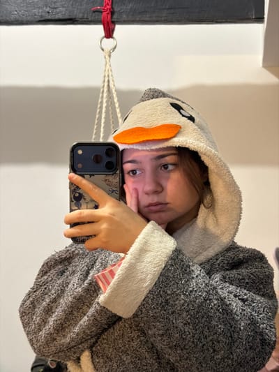 A person in a hooded, animal-themed onesie takes a selfie. The onesie is primarily grey and white. The person rests their head on a hand, looking into the camera. They hold a smartphone in front of them. In the background, there's a white wall, a dark beam with a red tie, and a hanging woven object. The image is vertically oriented.