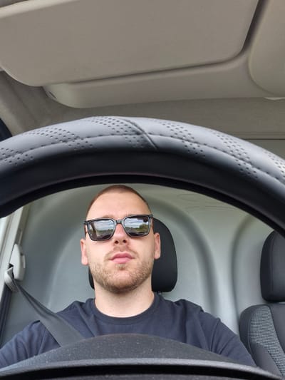 A man wearing sunglasses and a dark blue long-sleeved shirt is driving a vehicle. He is wearing a seatbelt. The steering wheel is light gray and has a perforated design. The interior of the vehicle is primarily gray. The scene cannot be identified as Graf Ignatievo, Bulgaria from the visual information provided.