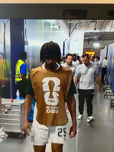 The image shows a person wearing a tan vest with "FIFA WORLD CUP 2026" printed on it, walking in a hallway. The person has dark, dreadlocked hair and is wearing white shorts with the number "25" printed on them. They are walking past other people in the hallway, some of whom are wearing official-looking attire. The hallway appears to be inside a stadium or sports facility. There are metal lockers or carts on the left side of the image, and people are walking in both directions. The environment is indoors with artificial lighting. No specific location in the city of Valencia, Spain is discernible from the image.