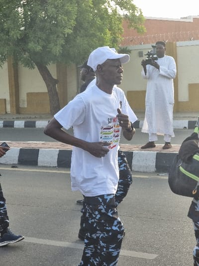 The image shows a man in a white t-shirt and a white cap running down a street in Maiduguri, Nigeria. He is wearing blue and black camouflage pants. He is running with his right arm bent, and he appears to be holding something with his left hand. Behind him, a person in camouflage pants walks, and another man wearing a white robe and a white cap appears to be filming the scene. In the background, a building with a yellow wall and a black fence is visible. The scene is a sunny day, and the street is paved with black asphalt and bordered by a black and white curb. A tree is to the left of the image. The coordinates provided indicate the location is in Maiduguri, Nigeria.